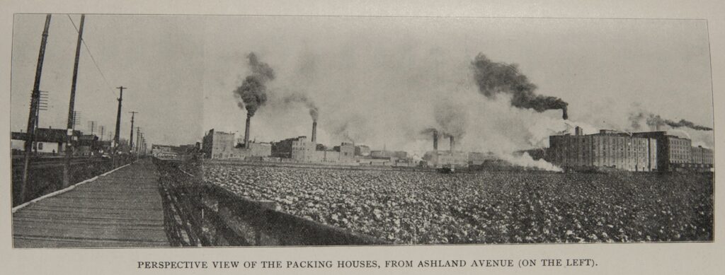Black-and-white photo of a wooden raised street on the right, with large stockyard in the foreground on the left. In the background are factories with smokestacks.