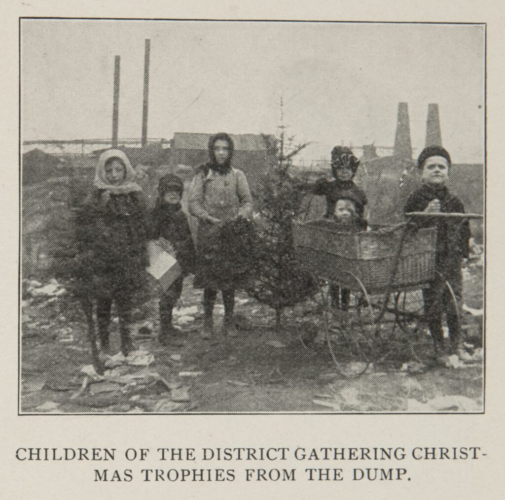 Black-and-white photo of six young children in threadbare winter clothes standing in a garbage dump. Two of them are holding small cut pine trees, while others hold boxes and a wreath.