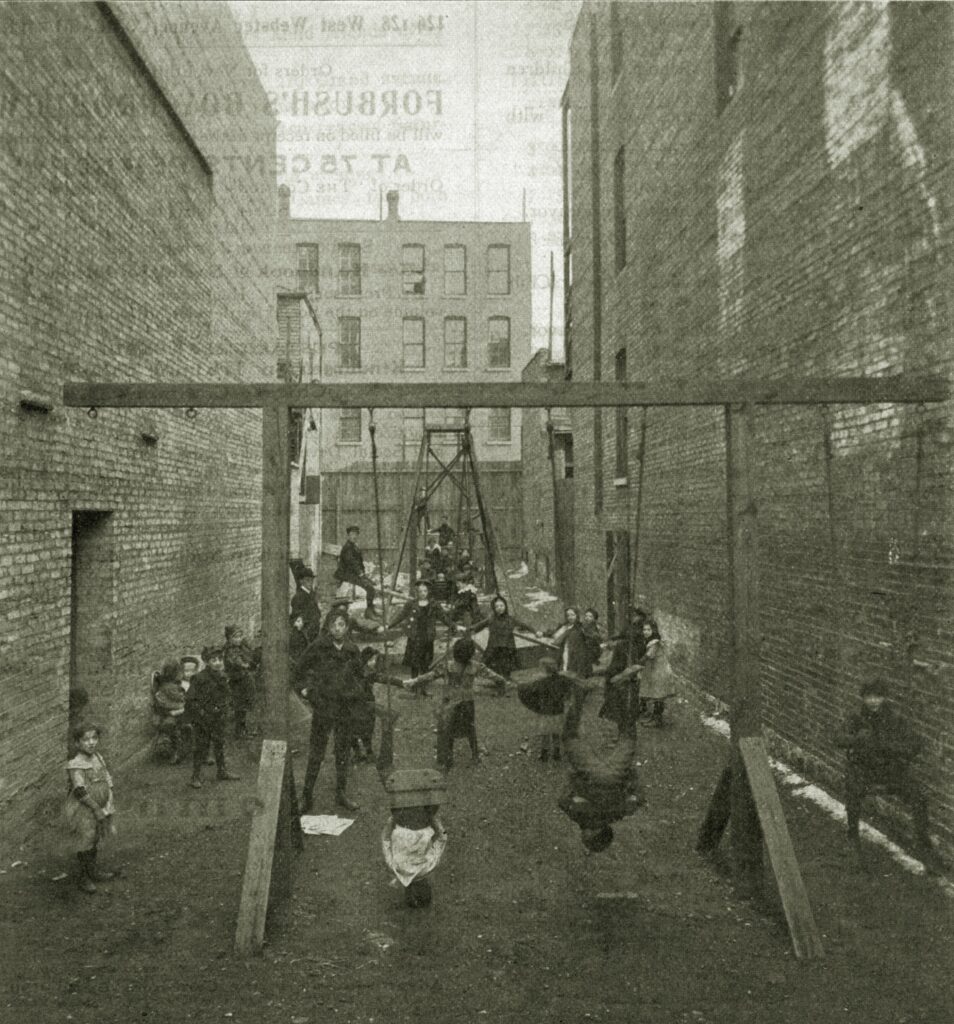 Black-and-white photograph of children playing in an unpaved alley between two apartment buildings.
