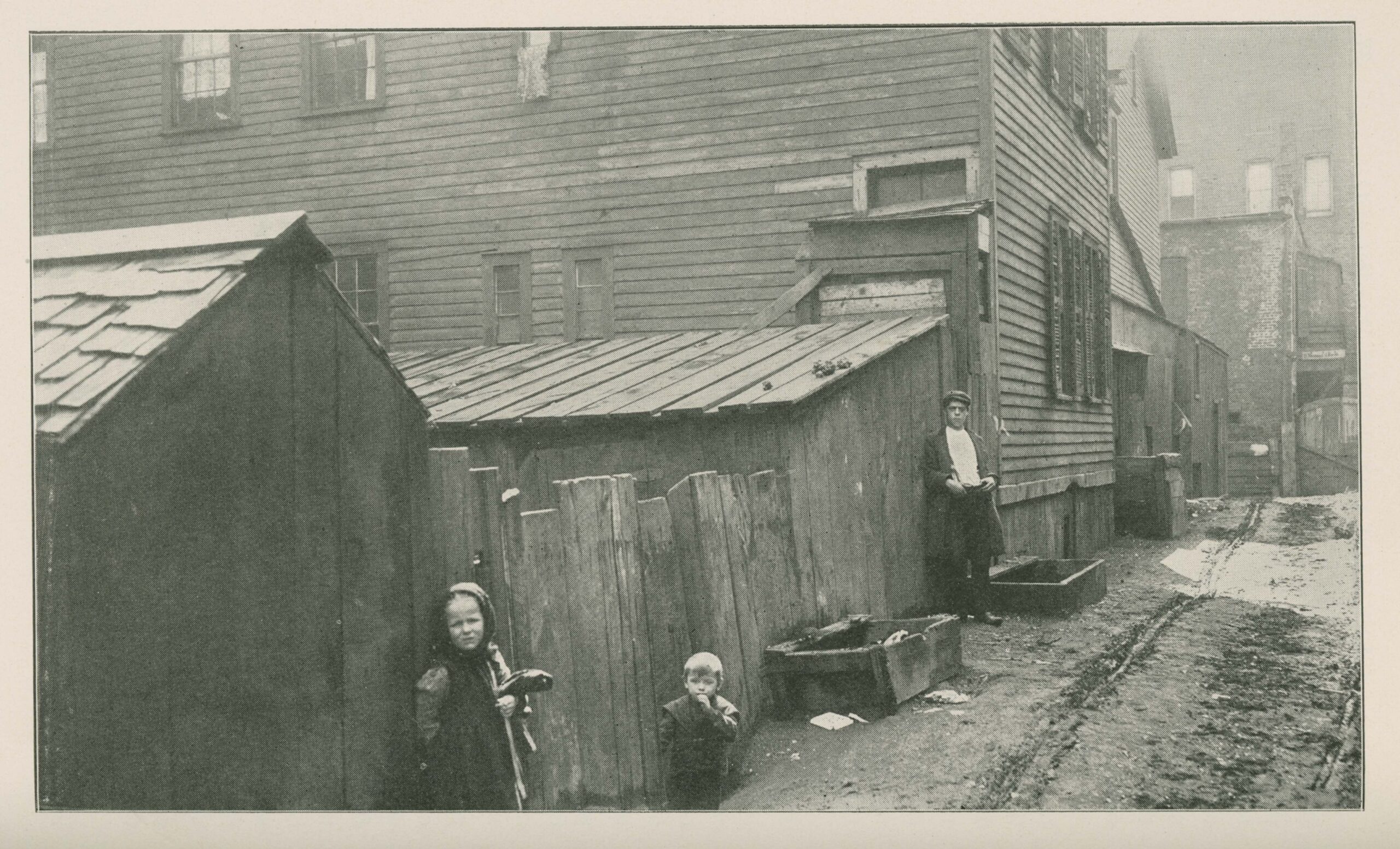 Black-and-white photo of a dirt street with houses and shacks on one side. A young child, a toddler, and a teenager stand at the edge of the road.