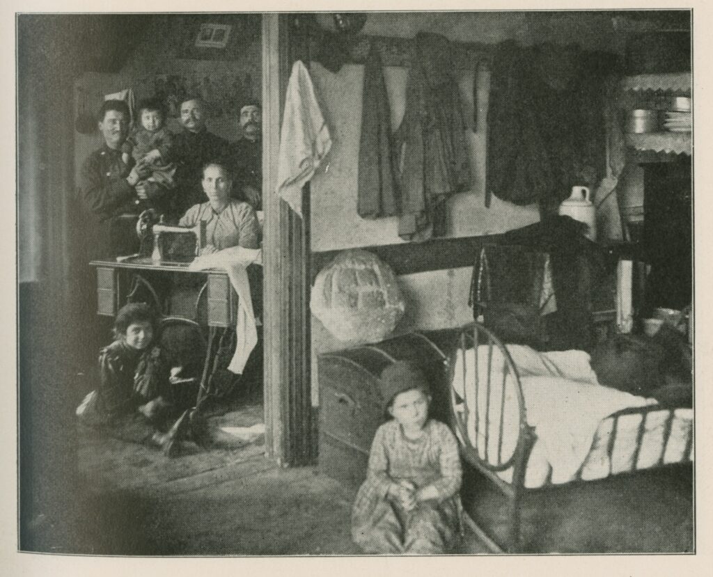 Black-and-white photo of the interior of a tenement house. In the foreground there is a small room with a crib, clothes hanging on the wall, and a small child sitting on the ground. In the background is another small room with a woman sitting a sewing machine with a girl sitting at her feet. Behind her are three men, one of whom is holding a small child.