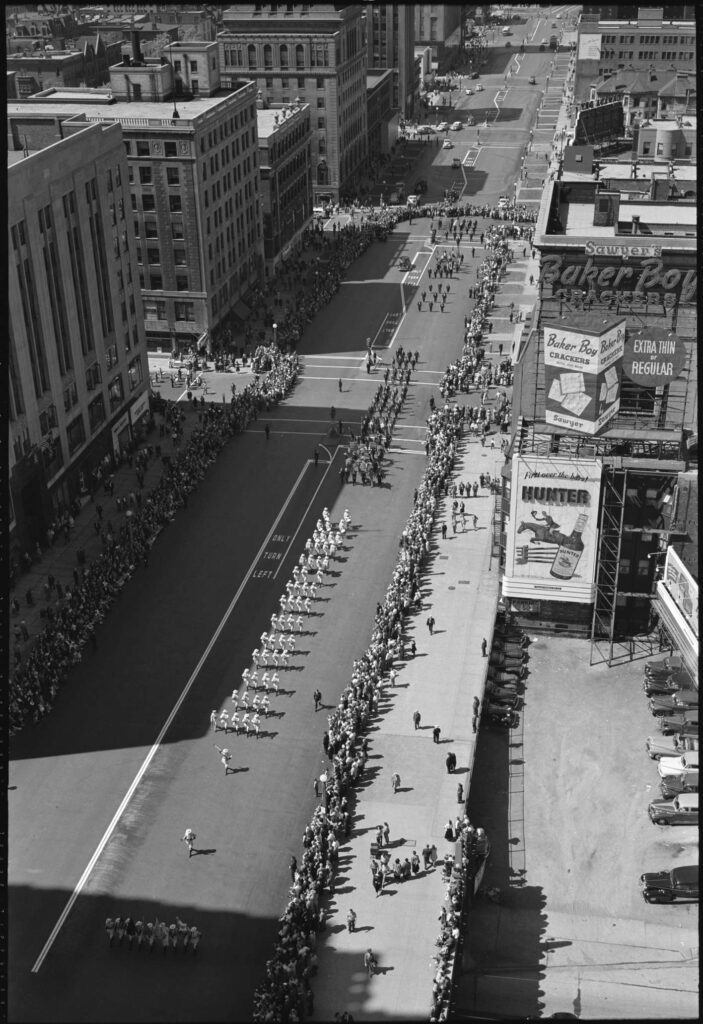 Black and white photograph of a parade running down a wide road between eight- and nine-story buildings. Buildings on the right-hand side of the street carry advertisements. People line the street three or four deep on both sides, while people in uniforms march three and four abreast down the middle of the street. Some carry flags and others carry instruments.