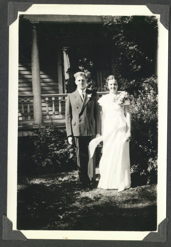 Black-and-white photograph of a white man and a white woman standing in a garden outside a house. The man wears a dark suit with a flower in the lapel and the woman wears a full-length white wedding dress with a bunch of flowers on her left shoulder. Both are smiling.