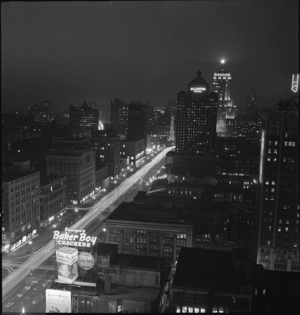 Black and white photograph of a nighttime city street scene. A wide street runs diagonally from the lower left to the middle of the image and intersects with another street in the lower left. Both streets are covered with lines of light (car headlights in a long exposure). Many eight and nine story buildings line the streets, with a few taller skyscrapers in the distance. Many windows in the buildings are light up, and some carry neon signs advertising foodstuffs and other things.