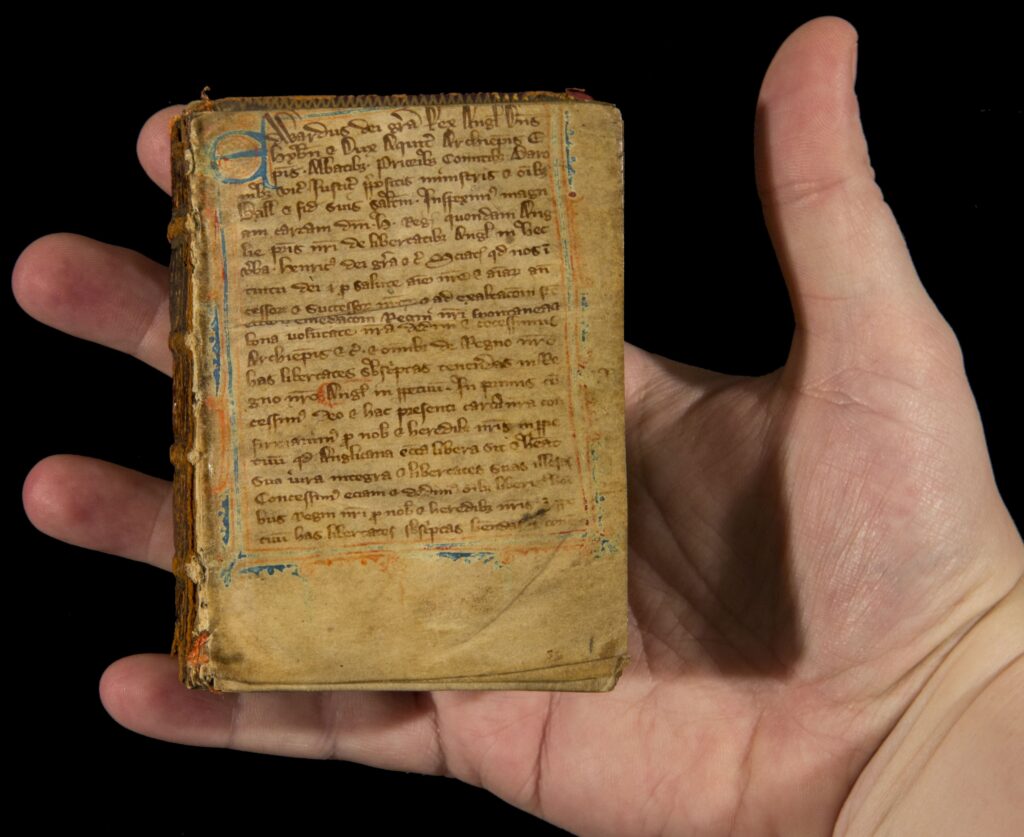 Image of a small medieval illuminated manuscript held in someone's hand so show that it is about the size of their plam. The page is three-quarters full of handwritten text, with a blue initial capital letter and a blue and red/orange border.