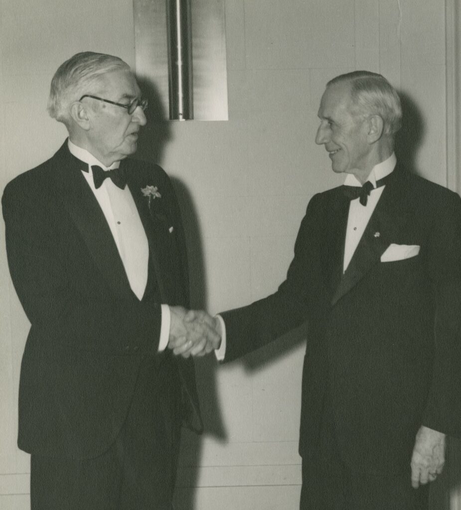 Black and white image of two white men wearing tuxedoes and shaking hands. The man on the left seems to be speaking while the man on the right smiles.