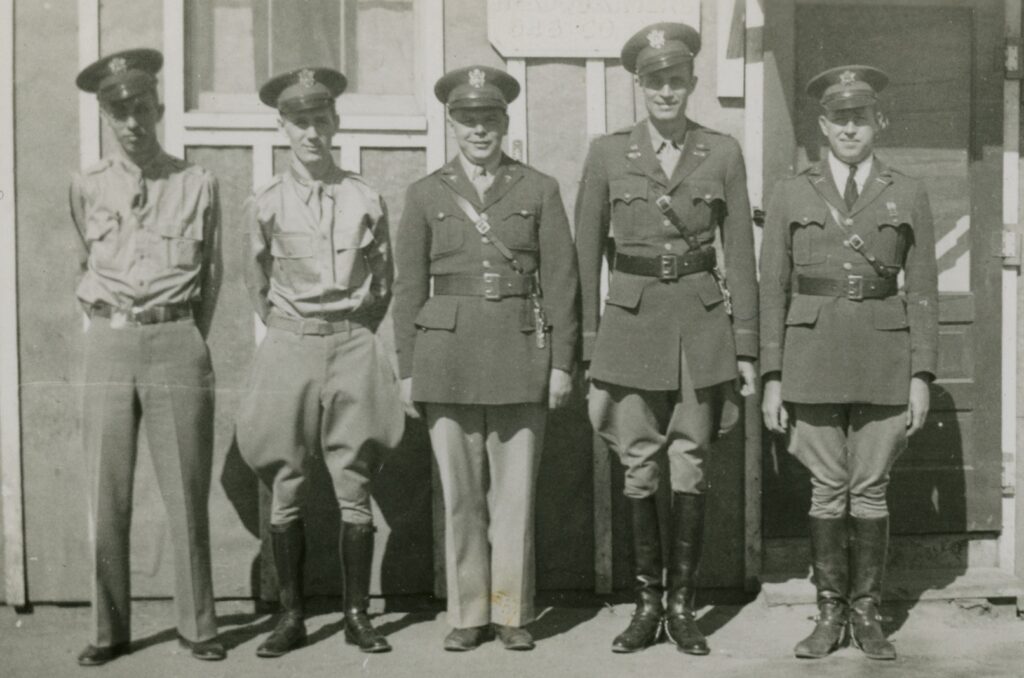 Black and white photograph of five white men in uniform standing shoulder to shoulder outside of a building.