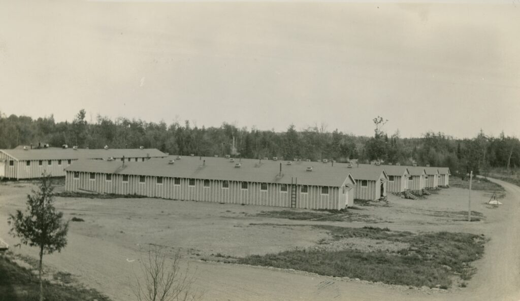Black and white photo of six long, one-story, pre-fab buildings standing in a row in a clearing. A seventh building stands behind the row of buildings, its long end facing their short ends. Dirt roads surround the buildings on two sides, and there are trees behind them.