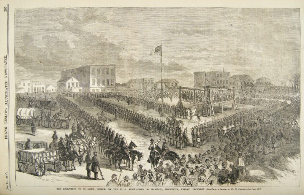 Engraving of the execution of 38 Dakota men by the US military. The men hang on a raised mass scaffold inn the middle-right of the image. They are surrounded by US soldiers in parade formation, with civilian spectators at the edge of the image. There are hundreds of people watching. There are buildings on the horizon, suggesting the execution happened in a town square or pubic place.
