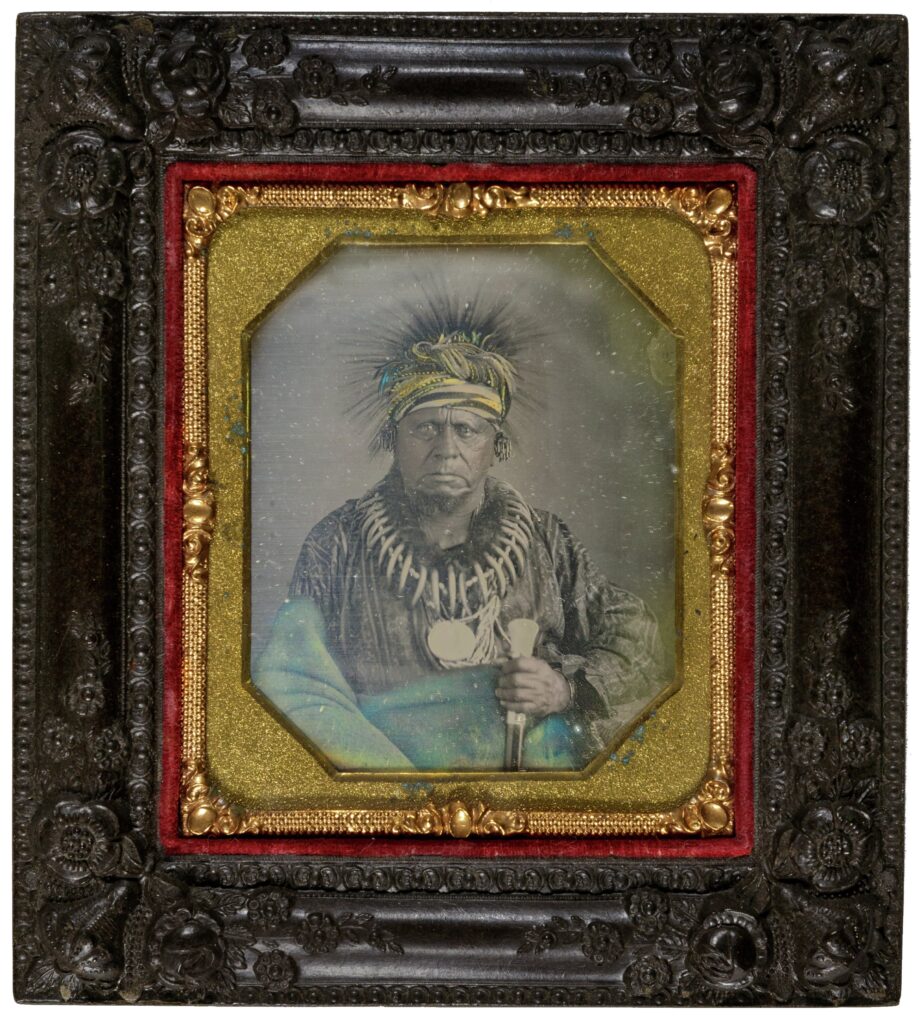 Black-and-white image in an ornate black-and-gold frame. Ke-o-kuk is an older man with dark skin and a serious expression. He wears a patterend shirt with a necklace of beads and animal claws and a large, central, circular medallion. Around his waist and right arm is a blanket. In his left hand he holds something--possibly a staff or gun--that extends down out of the frame. He wears a striped cloth wrapped around his head. Coming out of the top of this headdress are many fine lines, possibly his hair or fine animal quills. They make a star-like frame around his head.