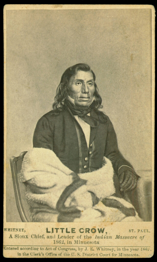 Black-and-white photo on top of a brief description. Che-tan-wa-ku-te-a-ma-na (Little Crow) is an older man with dark skin, a lined face, and shoulder-length dark hair. He wears a western-style suit and sits in a chair. A blanket is wraped around his waist and right arm, hiding it from view. His left arm rests on a table.