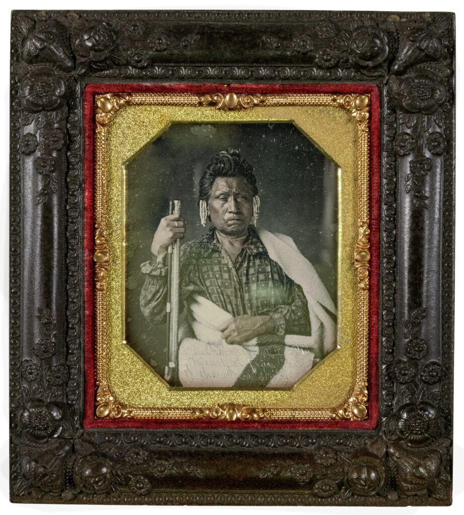 Black-and-white daguerreotype in an ornate black-and-gold frame. Na-che-ninga is an older, dark-skinned man with a serious expression. He wears a collared shirt and has a blanket wrapped over his midsection and one shoulder. He has long dark hair, which seems to be piled on his head. By either ear he wears dangling white bead hair ornaments. In his right hand he holds the muzzle of a gun, the bottom of which is resting on the floor out of the frame