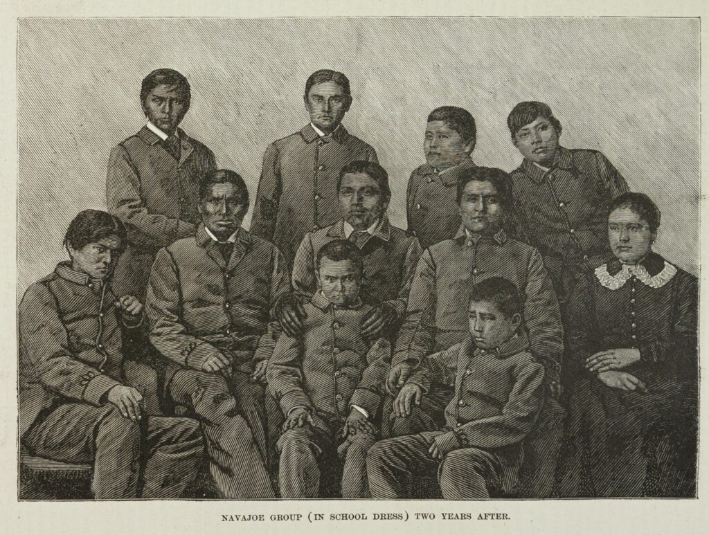 Black-and-white image of eleven Diné (Navajo) children in Western-style dress. Four stand in the back, five sit in the middle row, and the two youngest sit in front.