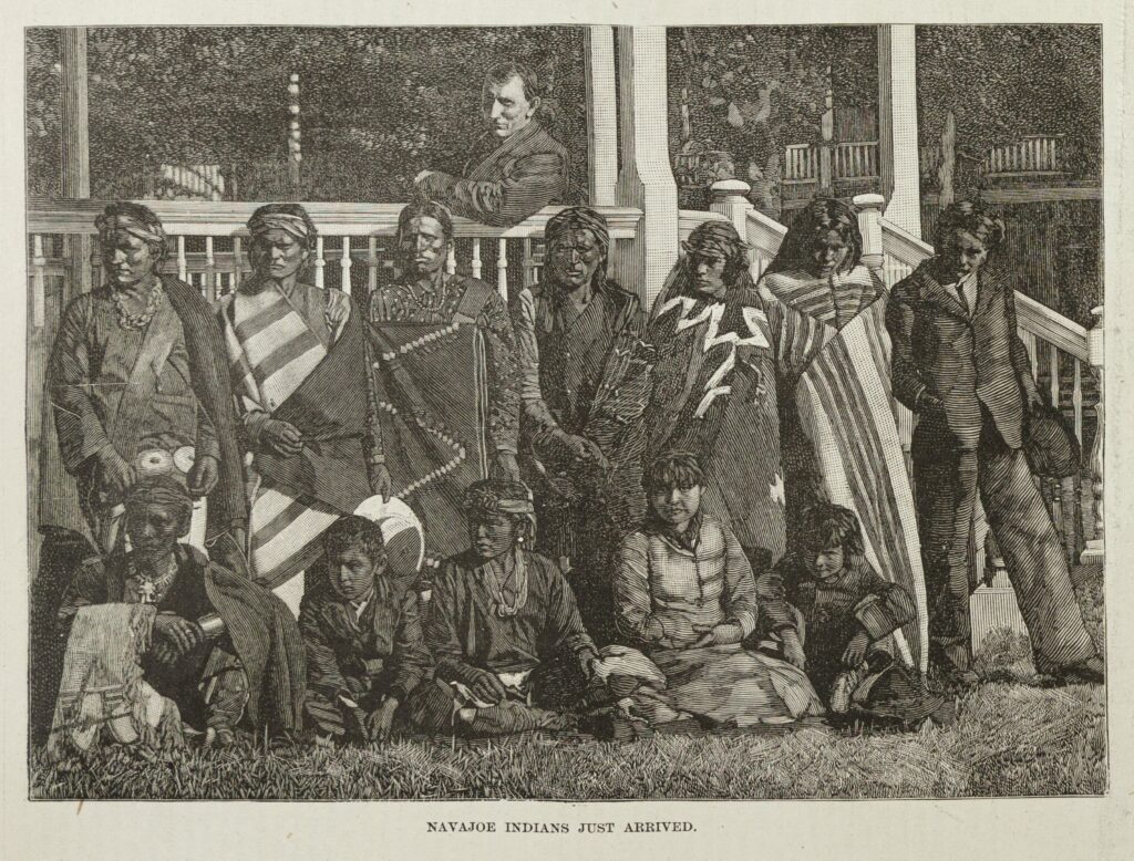Image of twelve Diné (Navajo) children wearing traditional clothing outside the front porch of a Western-style house. Seven children stand in the back row, five sit on the ground in front. A white man in a suit leans on the railing above them.