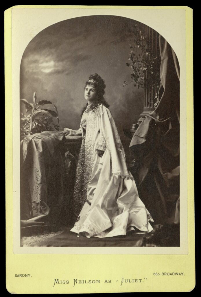 Black-and-white photo of Adelaide Neilson in costume as Juliet posed for the balcony scene. She wears a floor-length brocade dress and a long silk coat that trails in front of her. Her hair is partly piled on her head and partly down her back. She wears a small tiara.