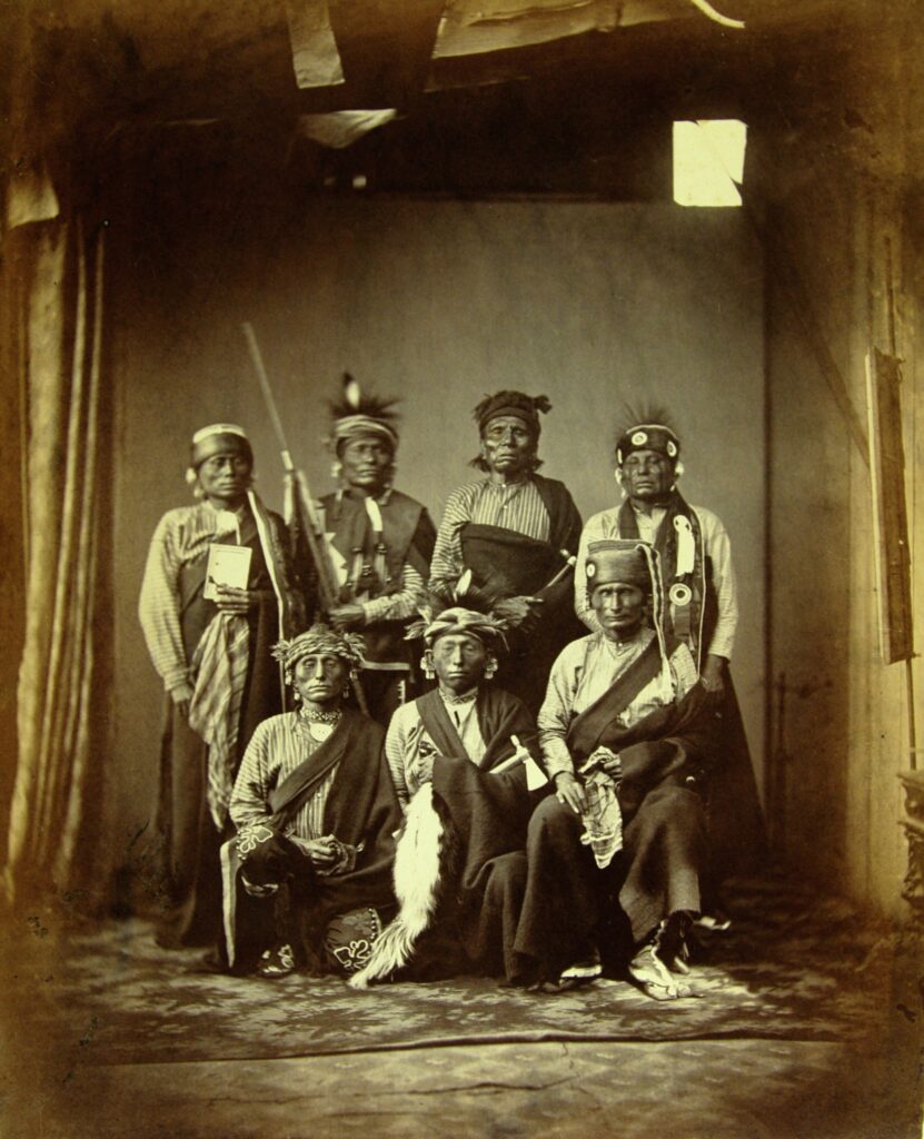 Black-and-white photo of a group of seven Indigenous men in traditional regalia posing for a studio photograph.