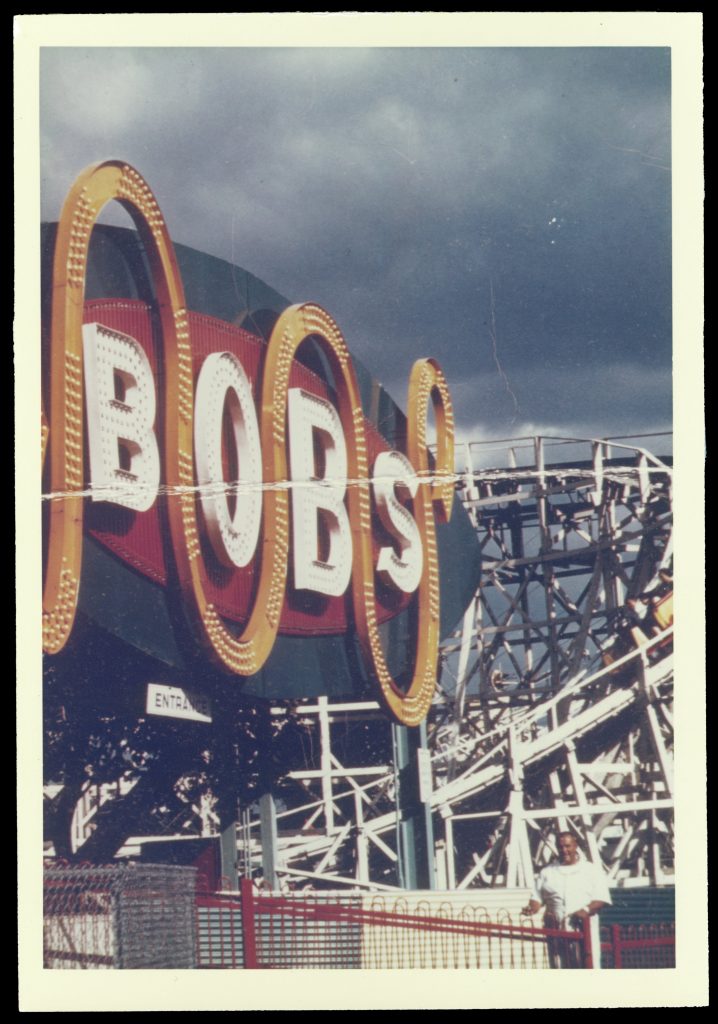 Color photograph of "The Bobs" ride sign. "Bobs" is written in white in all capitals, which an oranage line curving up and down between each letter. The letters and line have lightbulbs in them to illuminate them at night.