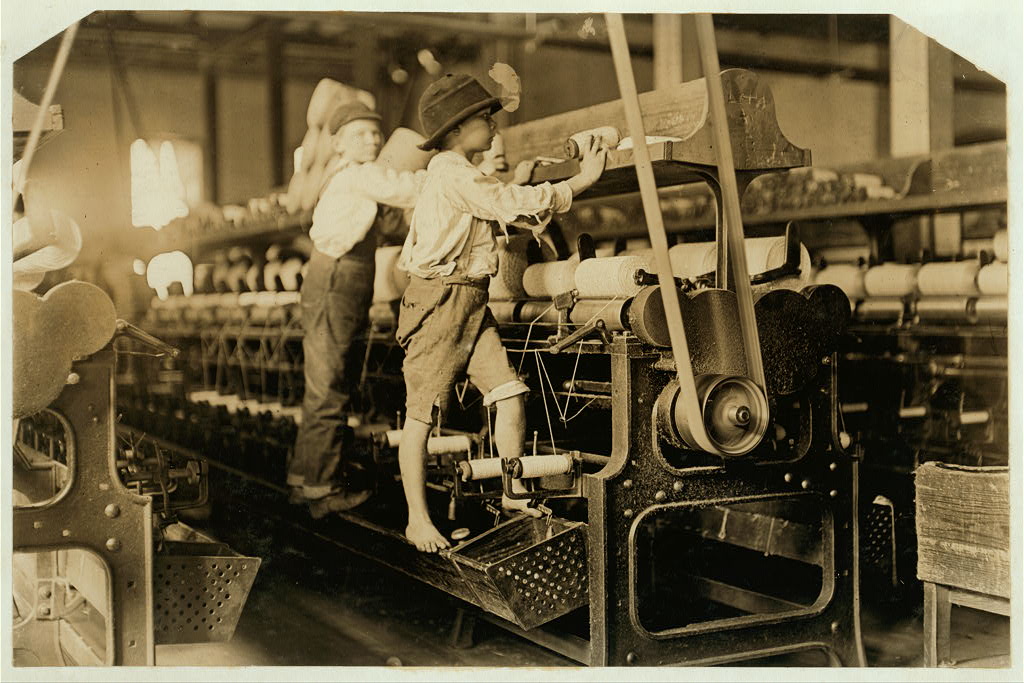 Sepia tone image of young boys working in a textile mill. They are barefoot and climbing on yarn winding machines.