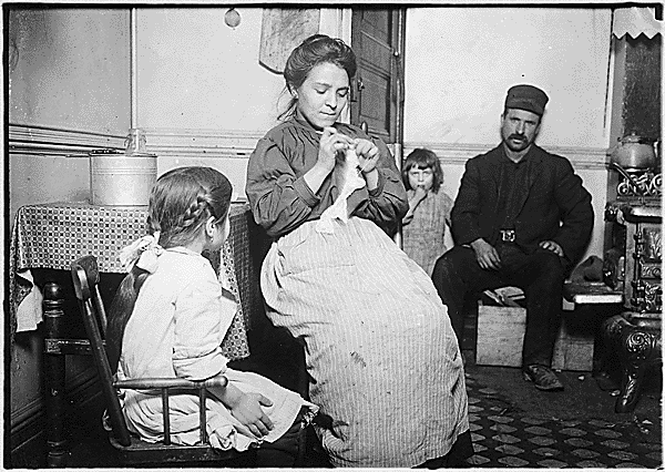 A woman in poor early 20th century dress sits in the middle of a scarcely-furnished room. She is making lace. Behind her on the viewer's right a man sits on a box and a small child stands sucking their fingers. An older girl sits in the left-hand foreground in a chair.
