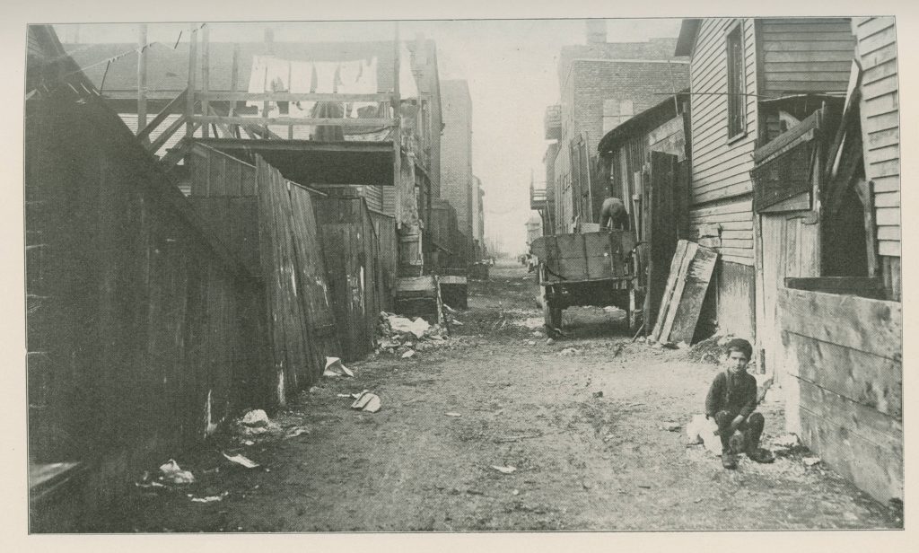 Black-and-white photo of a dirt street with shacks on either side. A young white boy sits on the ground in the right foreground.