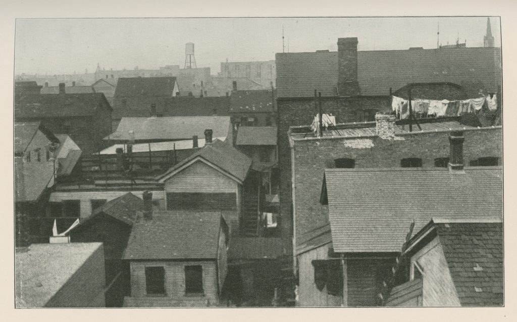Black-and-white photo looking across the backs of tightly packed one- and two-story houses. Laundry is hung across one of the rooves.
