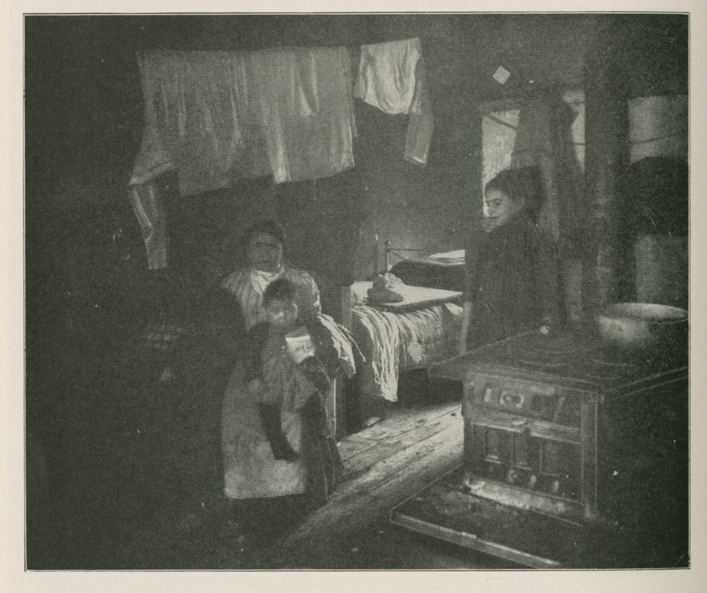 Black-and-white photo of a single small room with a bed, a woodstove, and laundry. A woman sits in the center with two children on her lap, and an older girl stands on the right.