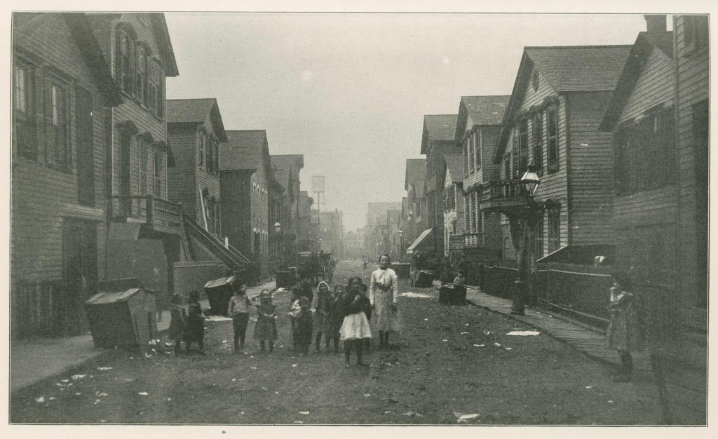 Black-and-white photo of a dirt street with two-story houses on either side. A group of children, mostly under 10, stand in the middle of the street.