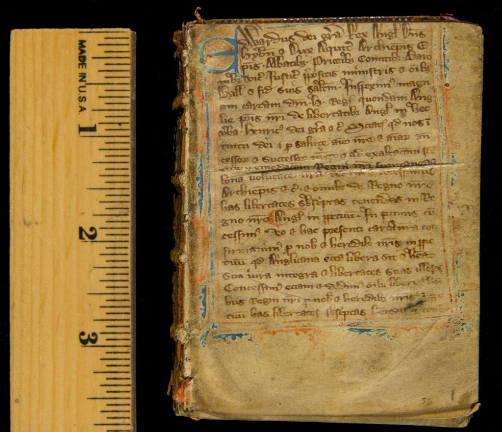 Image of a small medieval illuminated manuscript next to a ruler showing it is about 3.5 inches long. The page is three-quarters full of handwritten text, with a blue initial capital letter and a blue and red/orange border.