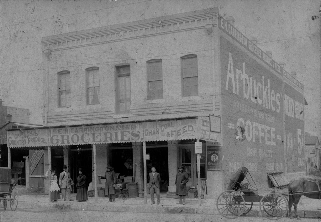 Black-and-white photograph of a two-story building standing alone and fronting a street with horse drawn vehicles. The first floor has a covered awning over the sidewalk labeled "E.H. Carrington Groceries" and has advertisements for a variety of products. Six well-dressed people stand under the awning.