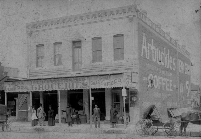 Black-and-white photograph of a two-story building standing alone and fronting a street with horse drawn vehicles. The first floor has a covered awning over the sidewalk labeled "E.H. Carrington Groceries" and has advertisements for a variety of products. Six well-dressed people stand under the awning.