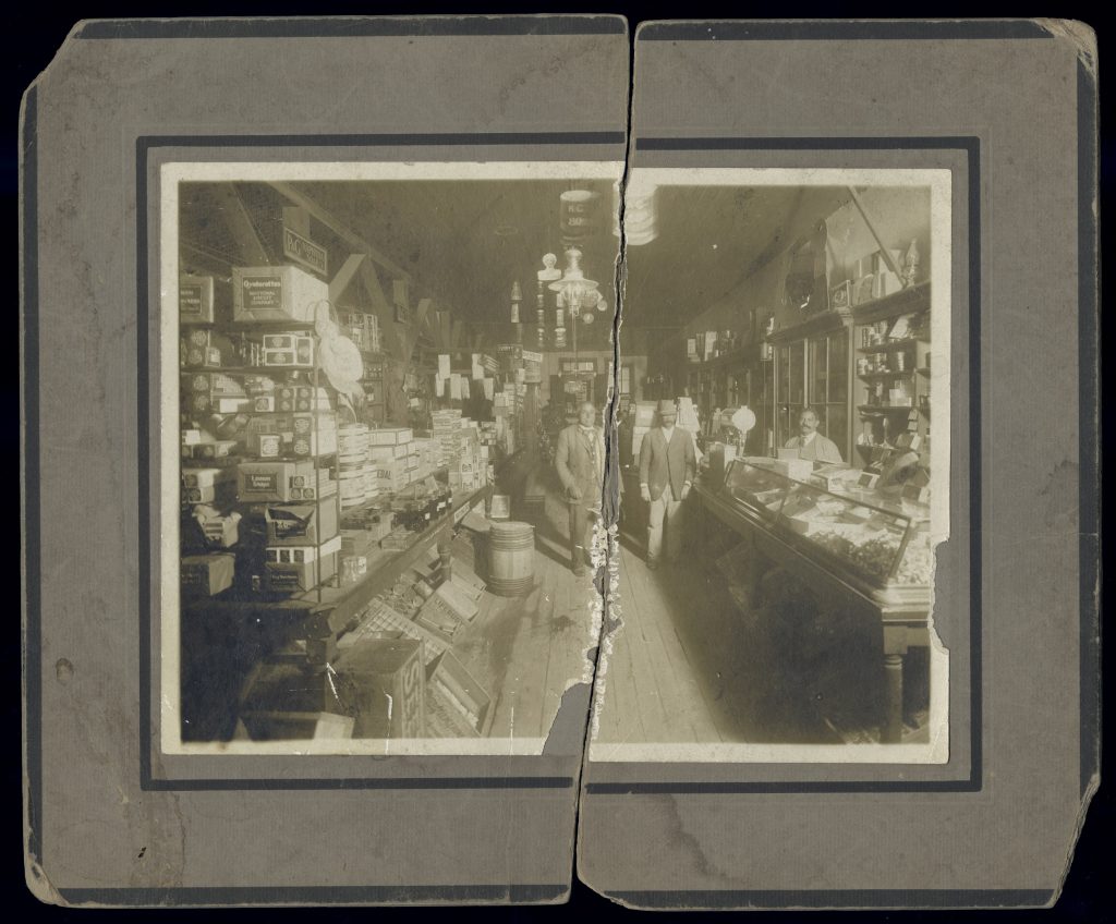 Black-and-white photo in a paper frame of the interior of a store filled floor to ceiling with products. Two counters line a narrow walkway in the middle of the store. Two Black men stand in the walkway, and another stands behind the counter on the right-hand side. The picture is broken down the middle.