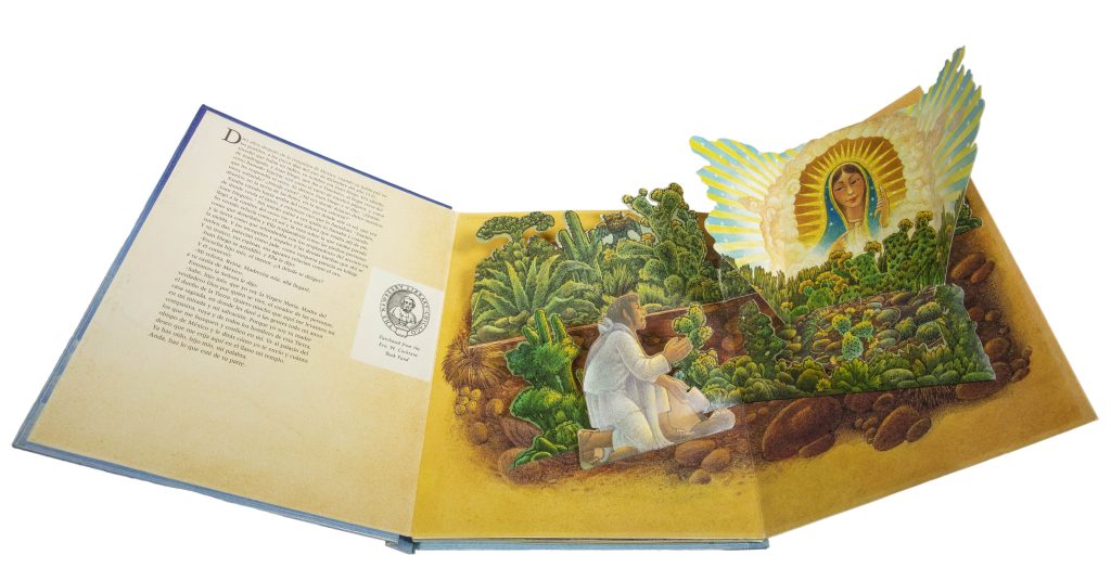 Book open to its first page, which folds out to create a pop-up image. In the image, a Latino man kneels in a landscape of rocks, cactuses, and other plants. In front of him the bust of the Virgin Mary appears surrounded by a halo of light and clouds. The man, some of the plants, and the Virgin Mary all pop out from the page.