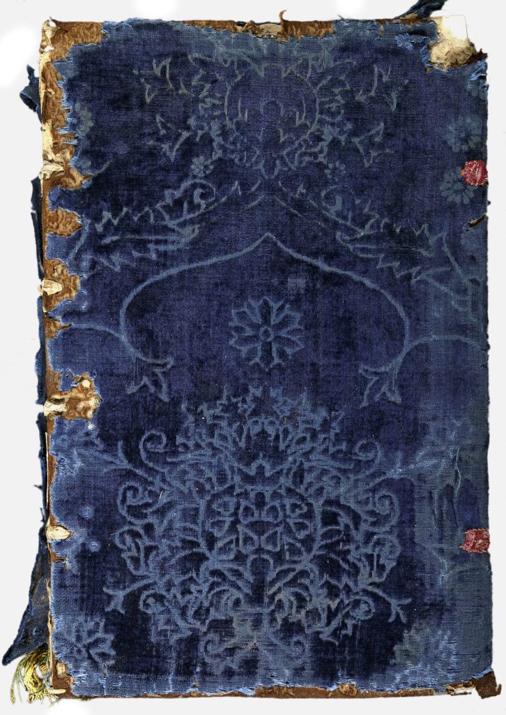 Cover of a book covered in blue velvet stamped with floral designs. The velvet has worn away on the corners and edge of the spine, revealing the board underneath.