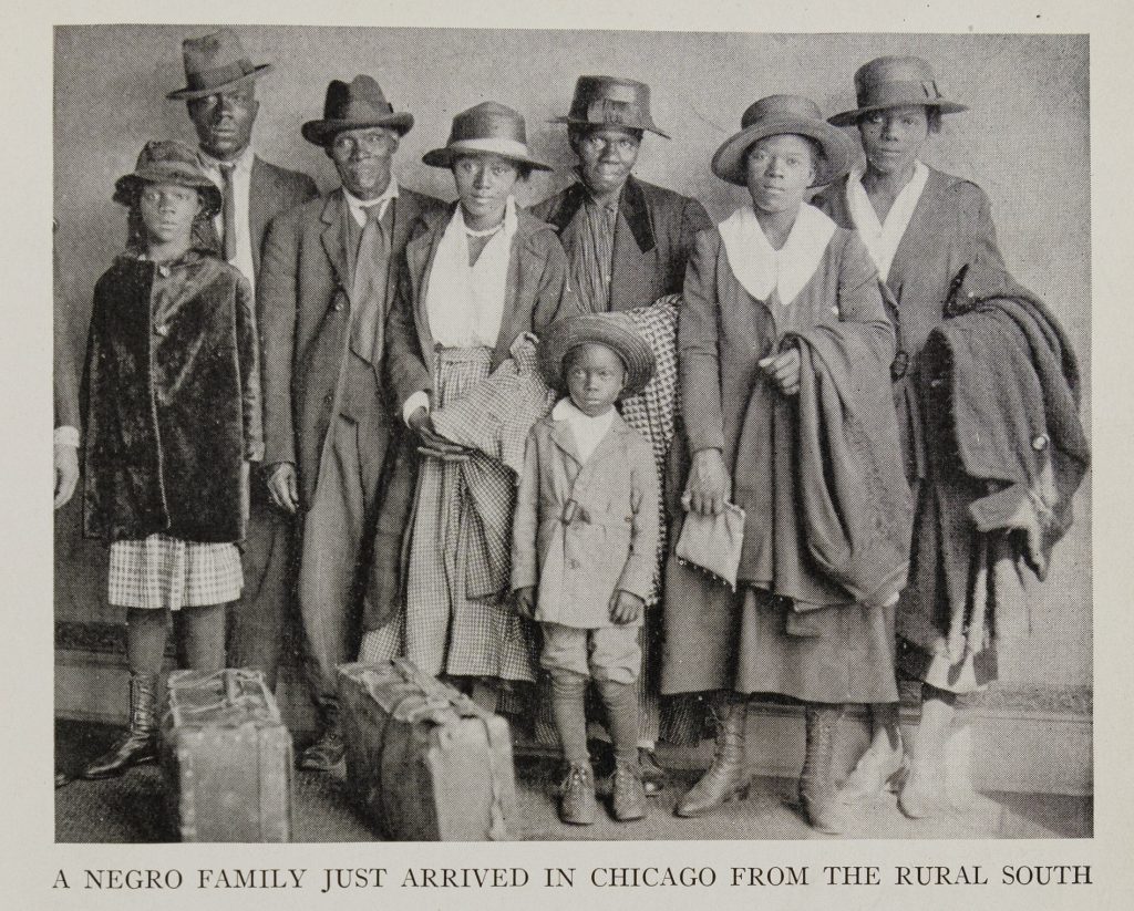 A black and white photo of a Black family (two men, four women, and two children) standing in a group. They wear coats and hats and have two suitcases in front of them.