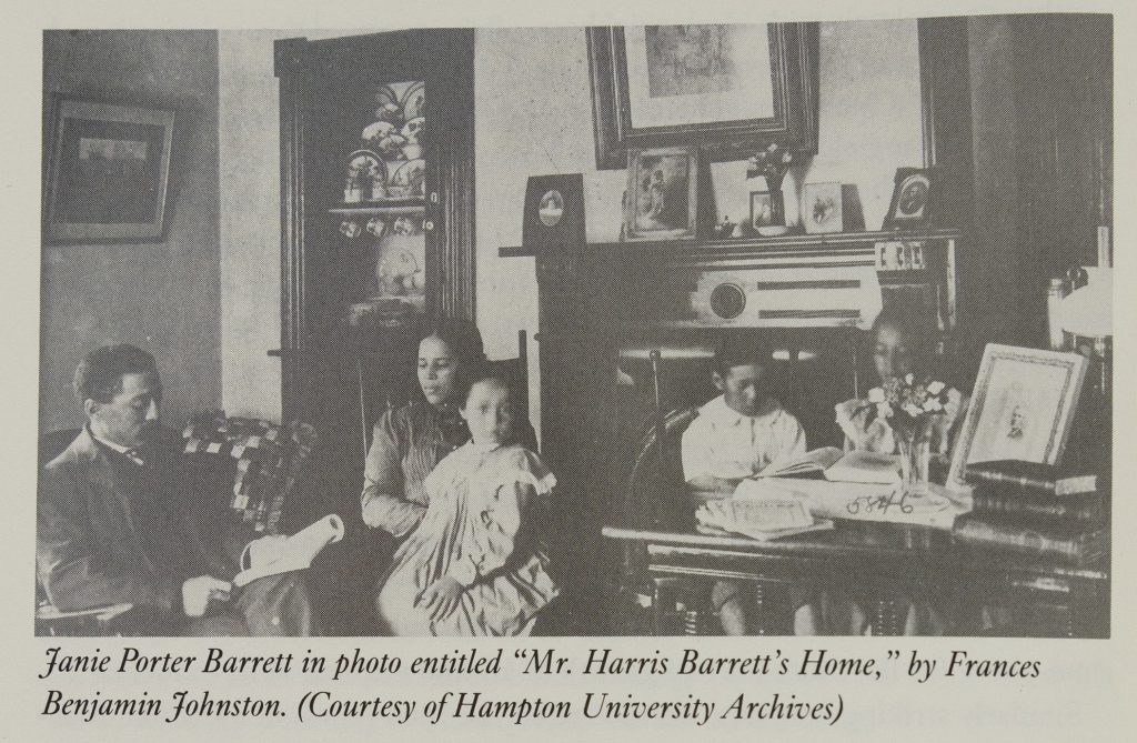 Black and white photo of Janie Porter Barrett and her family sitting in their living room. At the far left her husband sits reading a newspaper. Porter Barrett is next to him with a young child on her lap. Next to her are two older children sitting at a table. The caption reads, "Janie Porter Barrett in a photo entitled, 'Mr. Harris Barrett's Home,' by France Benajmin Johnston. (Courtesy of Hampton University Archives)."