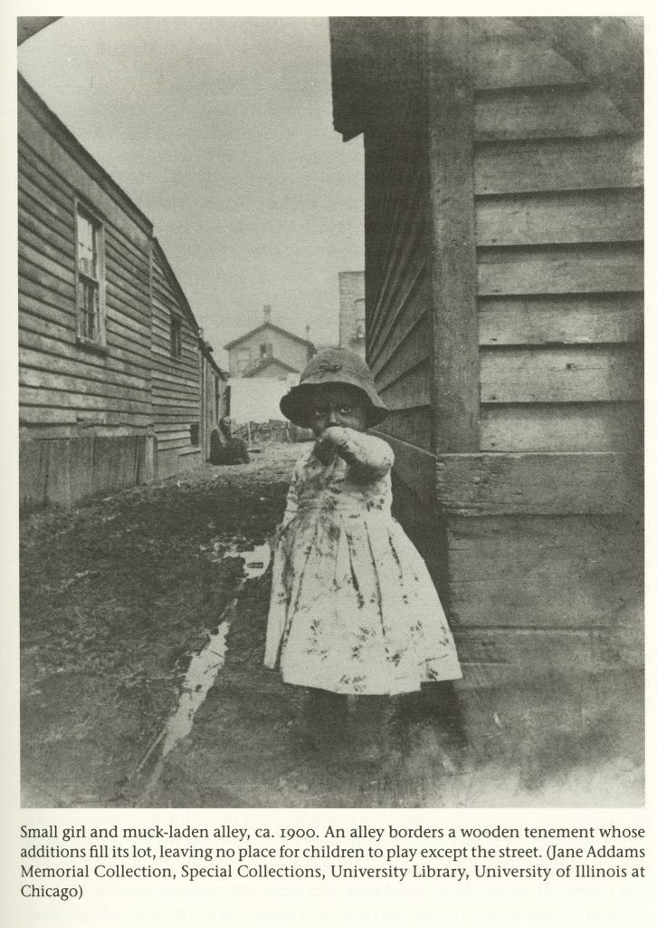 Black and white photo of a small girl (under five) standing in muddy alley between wooden single-story buildings. She wears a bowl hat and a light-colored dress. The caption reads, "Small girl and muck-laden alley, ca. 1900. An alley borders a wooden tenement whose additions fill its lot, leaving no place for children to play except the street. (Jane Addams Memorial Collection, Special Collections, University Library, University of Illinois at Chicago)."