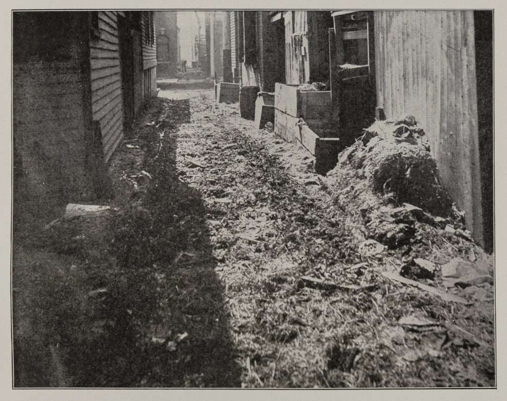 Black-and-white photo of an alley between two rows of wooden buildings. The alley is not paved and is a churned-up muck of debris and dirt.