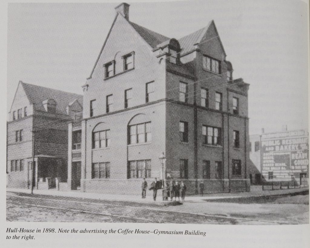 Black-and-white photograph of large four-story building sitting on the corner of two streets. A handful of people are standing in front of the corner where the streets meet. The photo's caption reads, "Hull-House in 1898. Note the advertising the Coffee House-Gymnasium Building to the right."