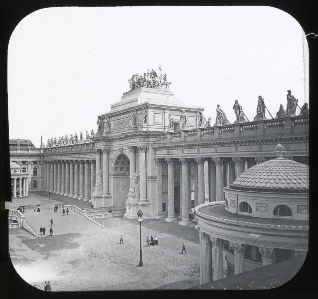 Black-and-white image of part of a courtyard bordered by an imposing two-story Greco-Roman colonnade of columns and statues. At the center one side of the colonnade is a triumphal arch topped by a statue of a horse-drawn chariot. People walk through the courtyard.
