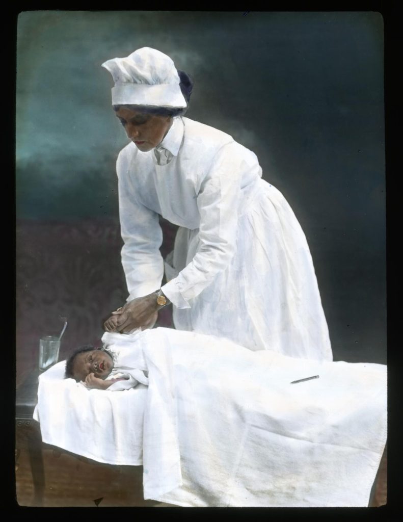 An African American women in a white nurse's uniform bends over an African American baby in a bed. She holds the baby's hand as if taking their pulse.