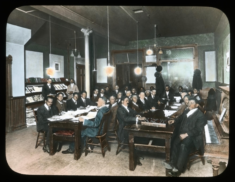 Approximately 30 Black men in suits and ties sit at two long tables in a small, well-decorated room. Most have reading material in front of them. Two stand at a rack of newspapers on the side of the room.
