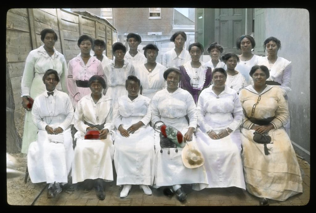 Eighteen well-dressed African American women posing for a picture. They sit and stand in three rows in an alleyway between a fence and a building.