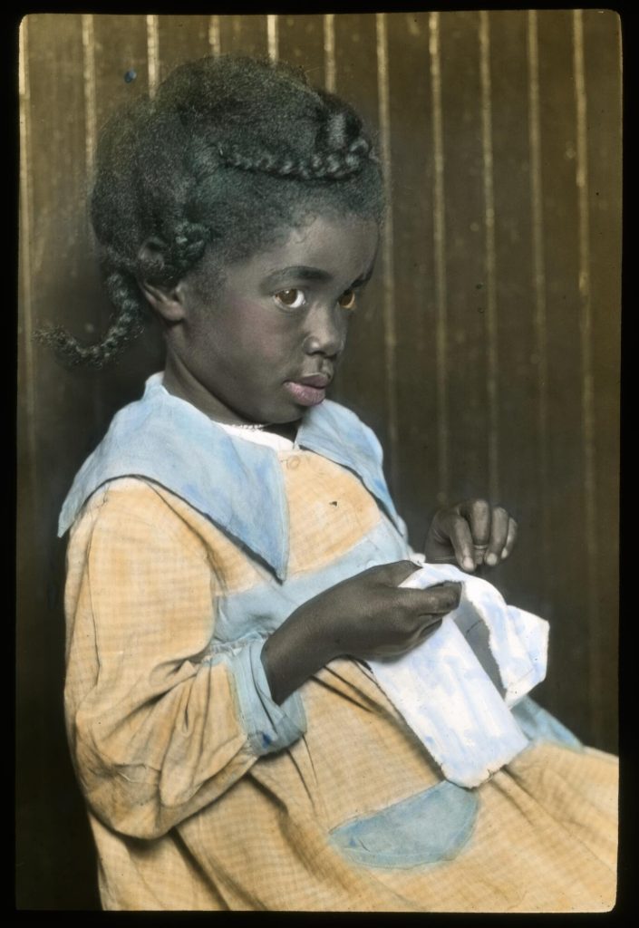Hand colored glass slide of a young African American girl sitting against a wall and hand sewing a small piece of fabric. She sits in profile and looks at the viewer out of the corner of one eye.