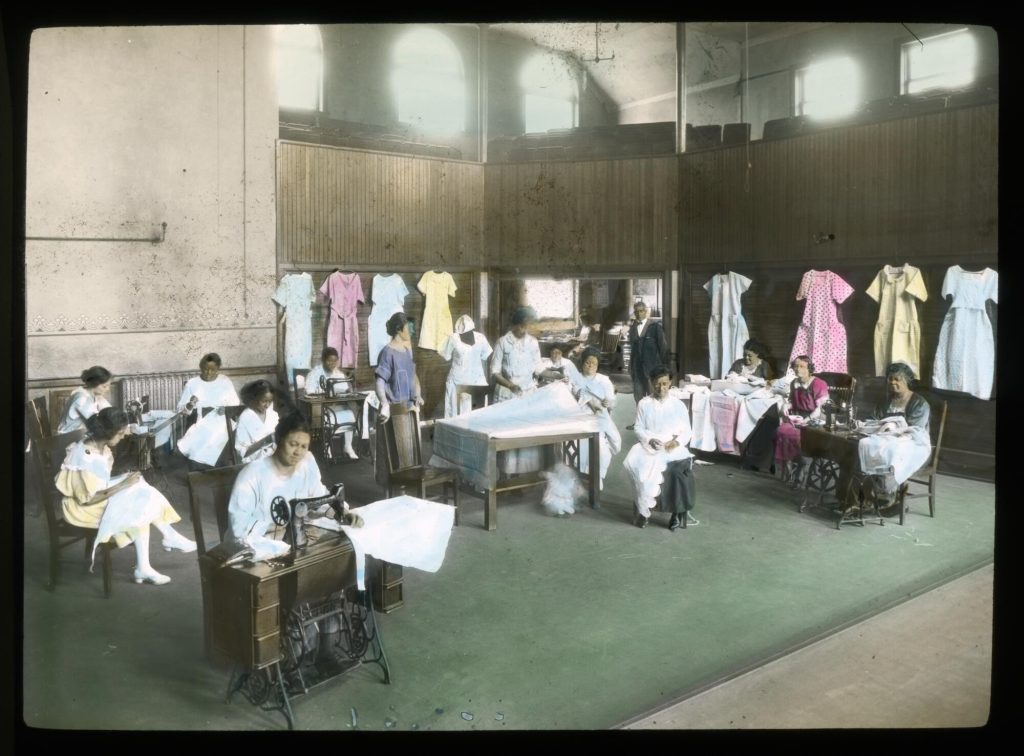 Colored glass slide showing 15 Black women working at sewing machines in a large hall. Completed dresses hang on the walls around them.