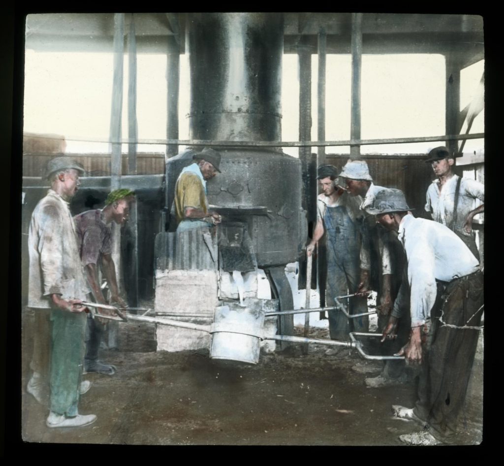 Hand-colored image of seven men collecting molten metal from a machine in a bucket. Five of the men are Black and two are white. All are wearing dirty, heavily-used clothes.