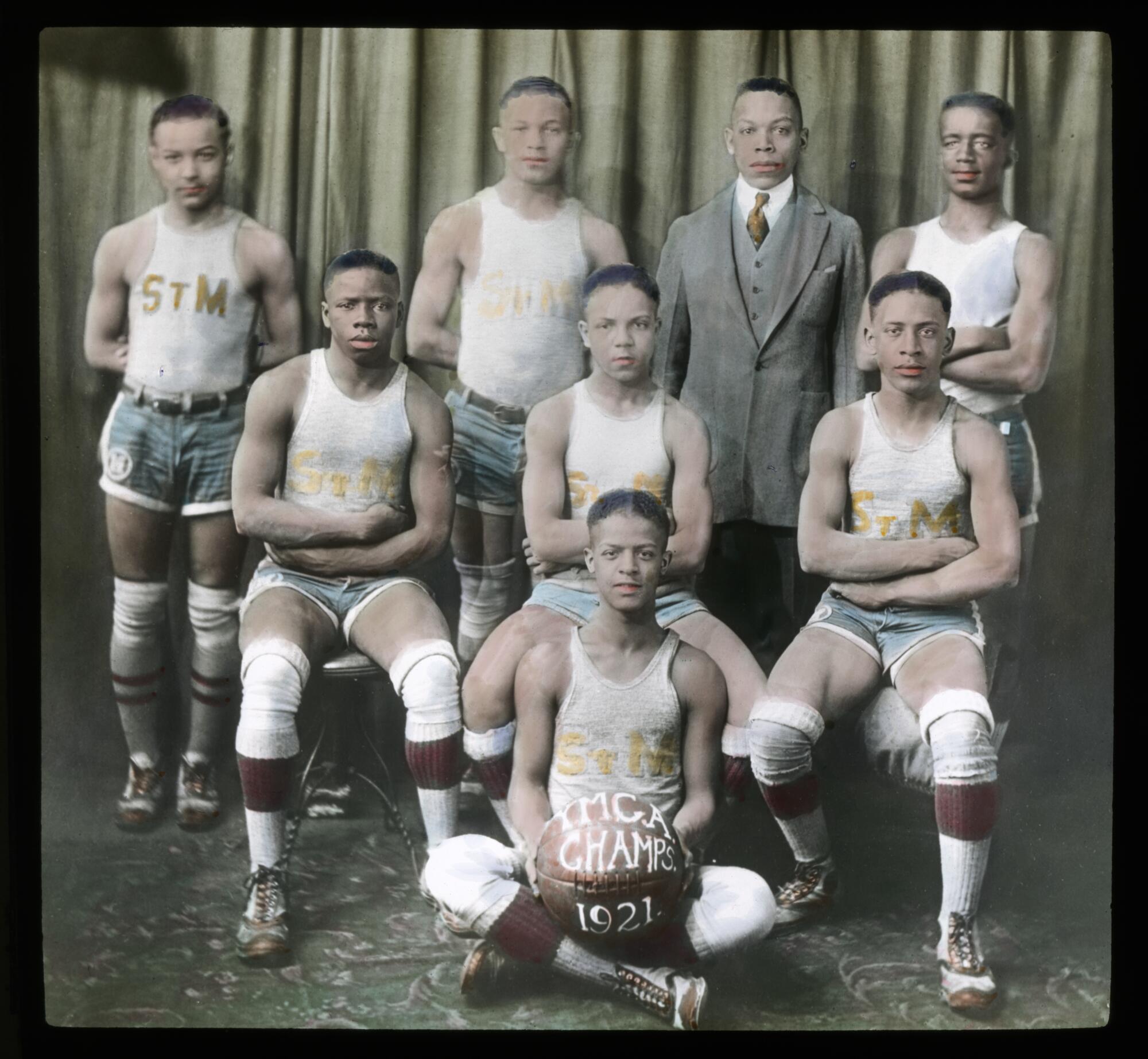 Hand-colored portrait of seven young Black men in basketball uniforms. Another Black man stands in the back row wearing a suit. The young man sitting in the front holds a basketball with the text "YMCA Champs 1921."