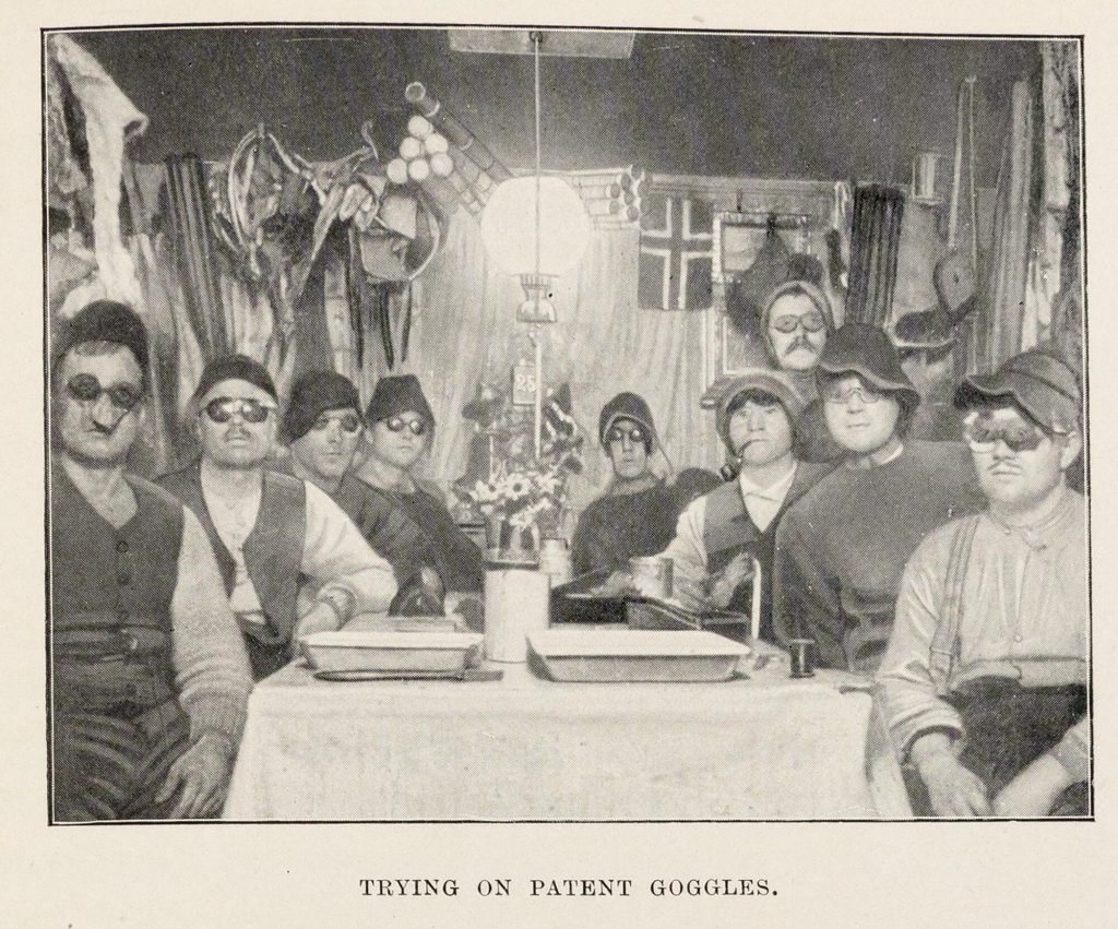 Black-and-white photograph of nine European men sitting around a table in a tent or small hut. Most are wearing dark goggles of various sizes.