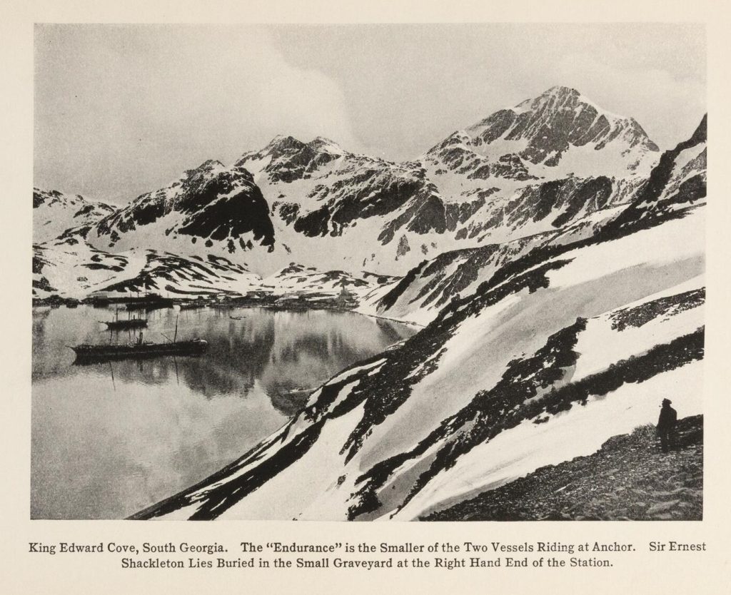 Black-and-white photograph of ships in a cove surrounded by tall. snow covered peaks.