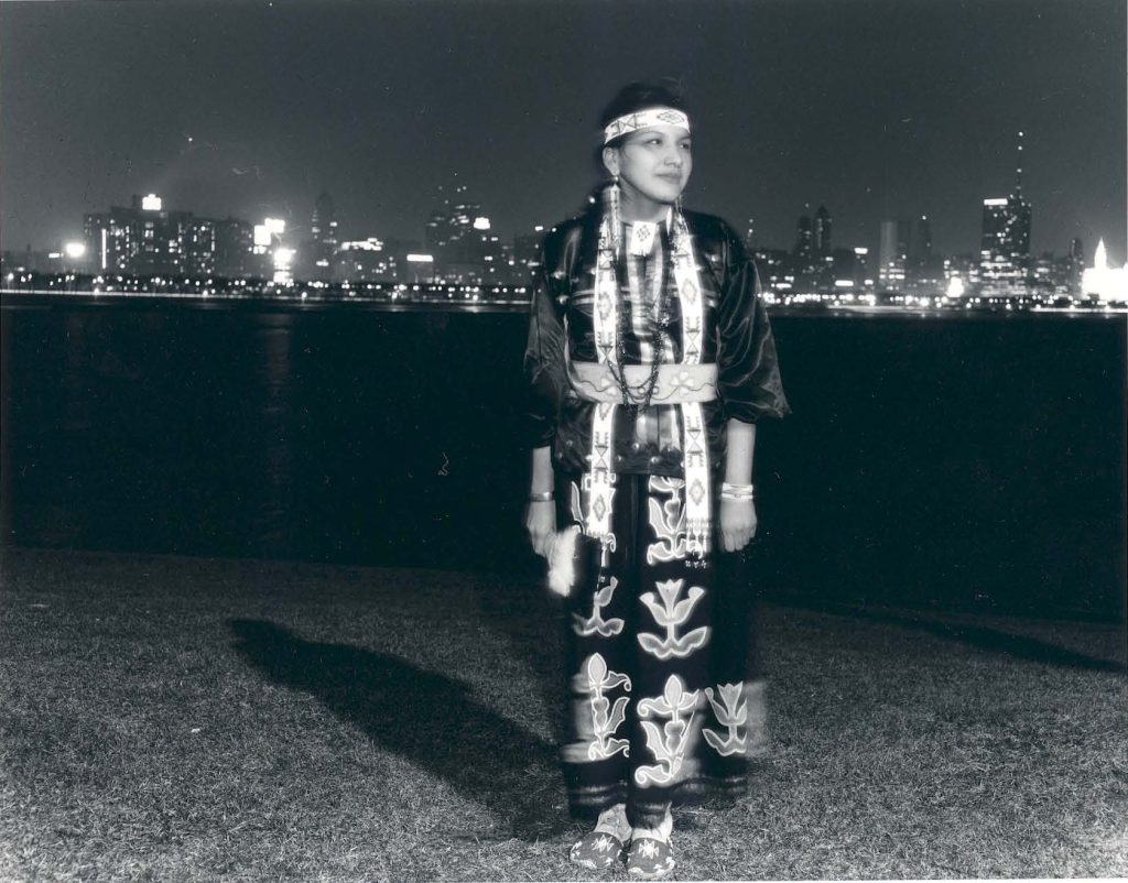 Balck-and-white photo of an Indigenous woman in regalia standing in front of the Chicago skyline lit up at night.