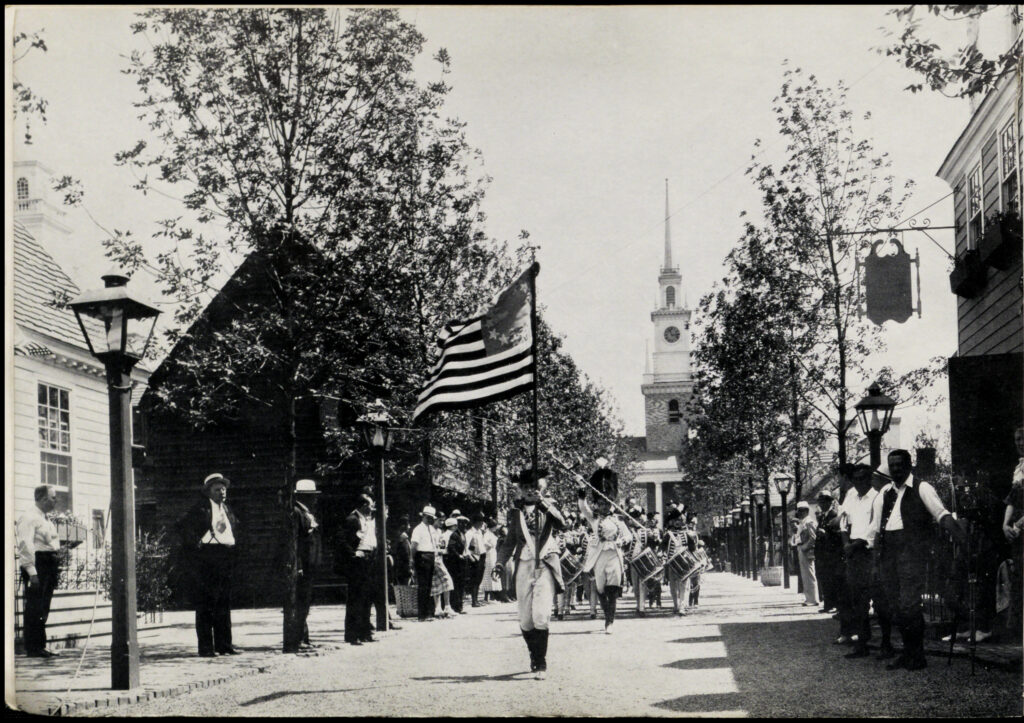 Black-and-white image of the Colonial Village at the Century of Progress. Spectators stand on a street lined with colonial-style houses as a marching band comes down the street. Members of the marching band are dressed in outfits influenced by the Continental Army, and the lead person carries the 13-star flag. Most, if not all, of the people in the picture are white.